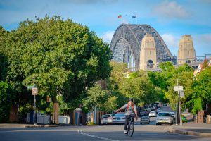 A cyclist rides down a street with Sydney Harbour Bridge in the background, surrounded by lush trees.