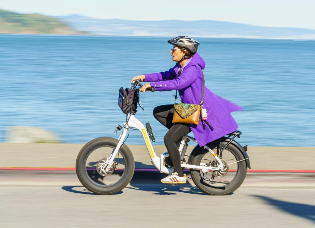 A woman rides an electric bicycle along the waterfront, wearing a purple coat and helmet, on a sunny day.